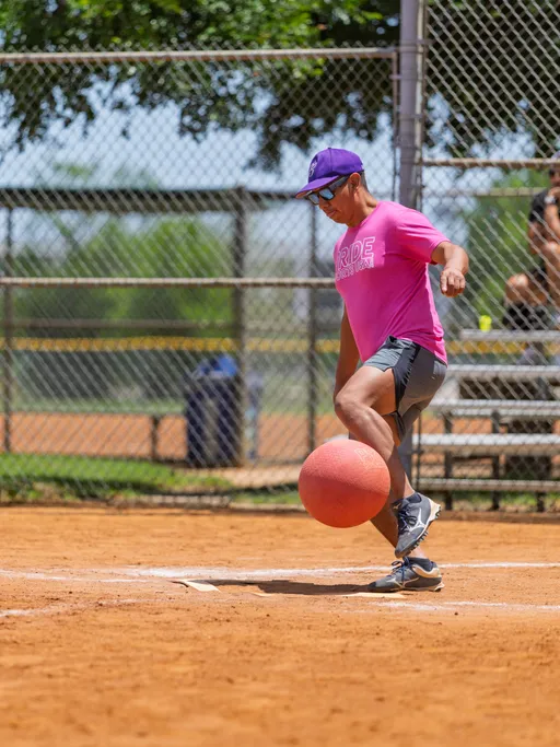 A player wearing a bright pink "PRIDE" shirt, sunglasses, a purple cap, and athletic shoes is kicking a red kickball on a dusty kickball field, with a chain-link fence and bleachers visible in the background.