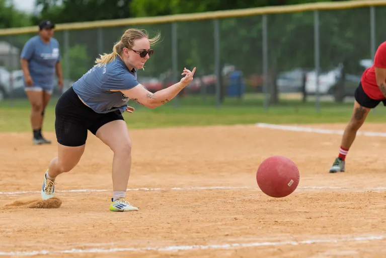 A player wearing a gray shirt, black shorts, and sunglasses is pitching a red kickball on a sandy field, while other players are positioned in the background and to the side, ready for action.