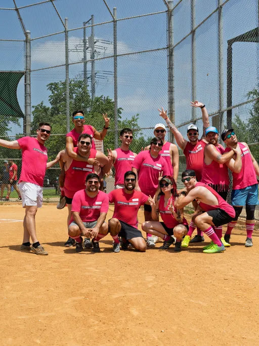 A group of kickball players wearing matching bright pink shirts and sunglasses pose playfully together on a dusty kickball field, with some crouching, some kneeling, and others giving piggyback rides or making celebratory gestures, surrounded by chain-link fencing and trees in the background.
