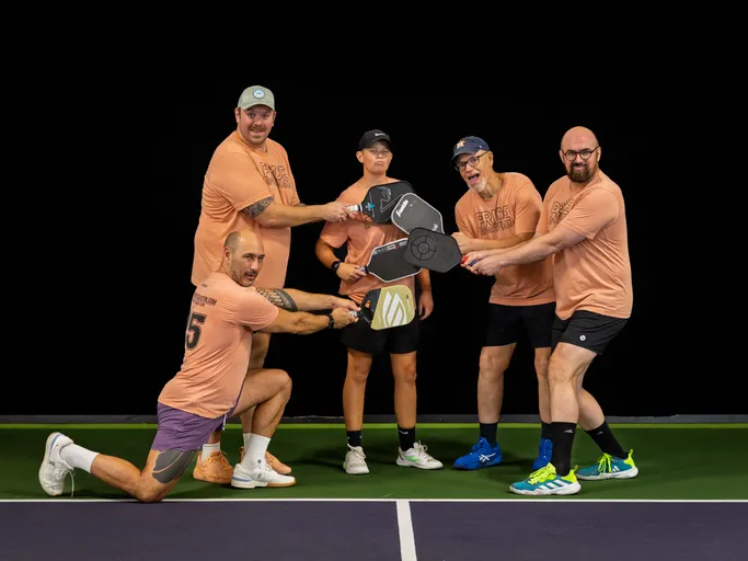 Five pickleball players pose together on an indoor court, all wearing matching peach-colored "Pride" shirts, shorts, and athletic shoes while playfully stacking their paddles at the center.