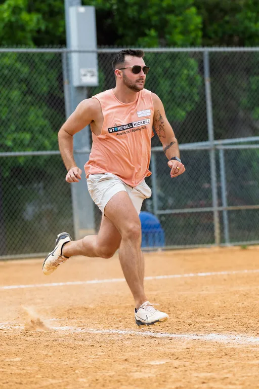 A kickball player in a sleeveless Stonewall Kickball shirt, shorts, athletic shoes, and sunglasses is running on a dirt field, with a chain-link fence and green trees in the background.