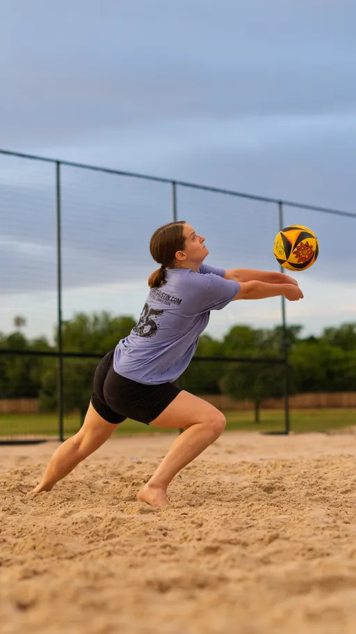 A volleyball player is lunging forward on a sand court with arms outstretched, preparing to bump a colorful volleyball in midair; the player is wearing a light purple shirt and black athletic shorts. A chain-link fence and trees are visible in the background.