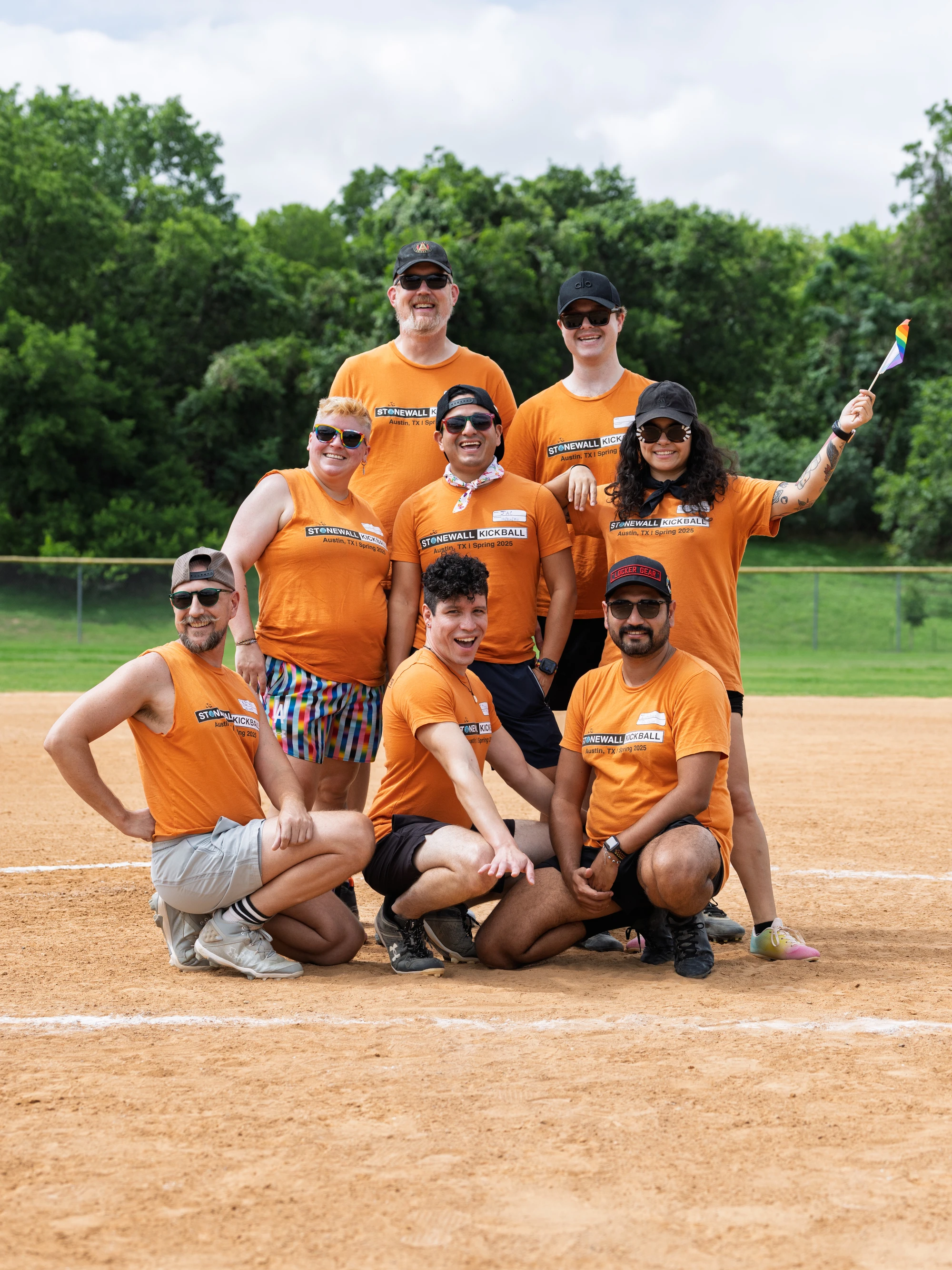 A group of kickball players wearing matching orange "Stonewall Kickball" shirts pose together on a kickball field, with some kneeling in front and others standing behind; one person is holding a small rainbow flag, and the background shows a grassy, tree-lined area.