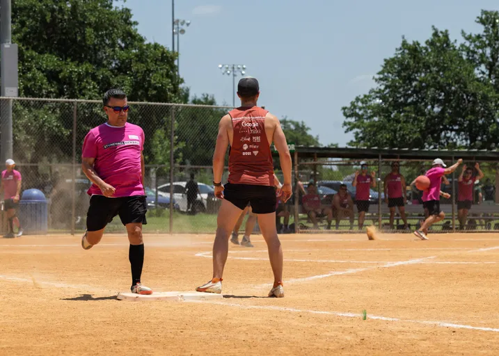 A player in a pink shirt and black shorts runs toward a base, while another competitor in an orange tank top stands near the base on a sandy kickball field, with more players and a kickball visible in the background.