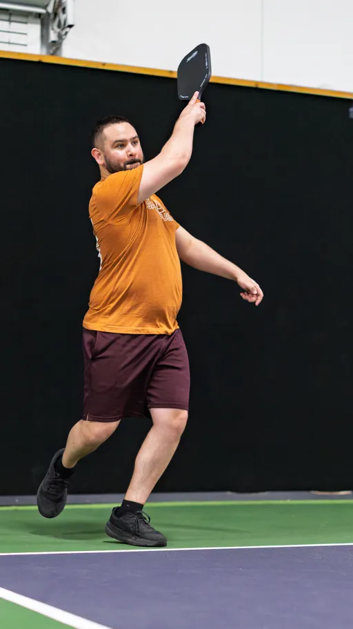 A pickleball player wearing an orange shirt, maroon shorts, and black athletic shoes is captured mid-action on an indoor court, swinging their paddle during a game.