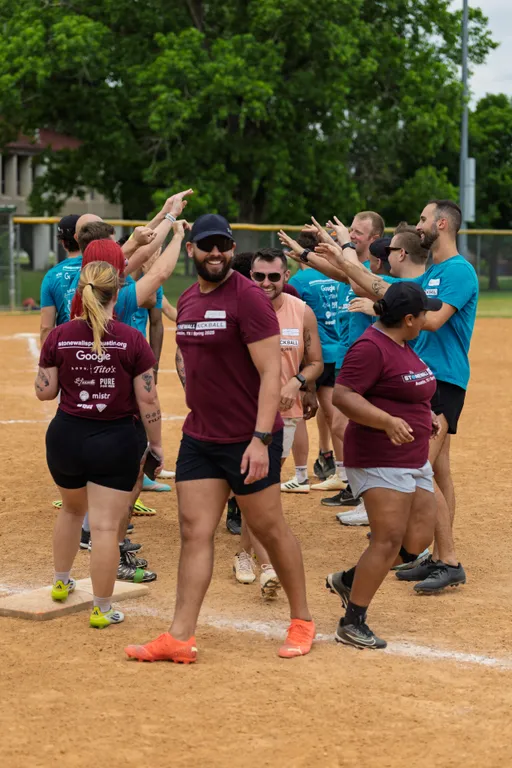 Players from two kickball teams, wearing maroon and teal shirts, line up and exchange high-fives on a dirt kickball field surrounded by grass and trees. Several players are wearing athletic shorts, hats, and sunglasses, and the atmosphere appears celebratory and friendly.
