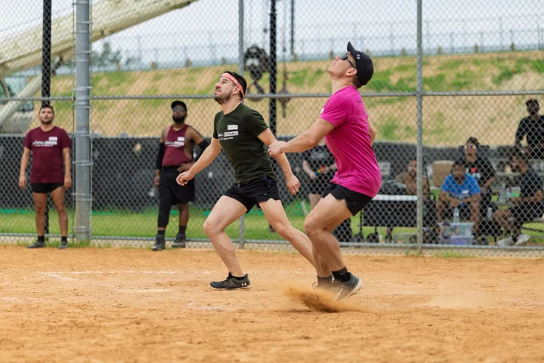Two kickball players wearing athletic shirts and shorts, one in green with a headband and the other in pink with a cap, are focused on the ball overhead while moving on a dirt field; additional players and spectators are visible near the fence in the background.