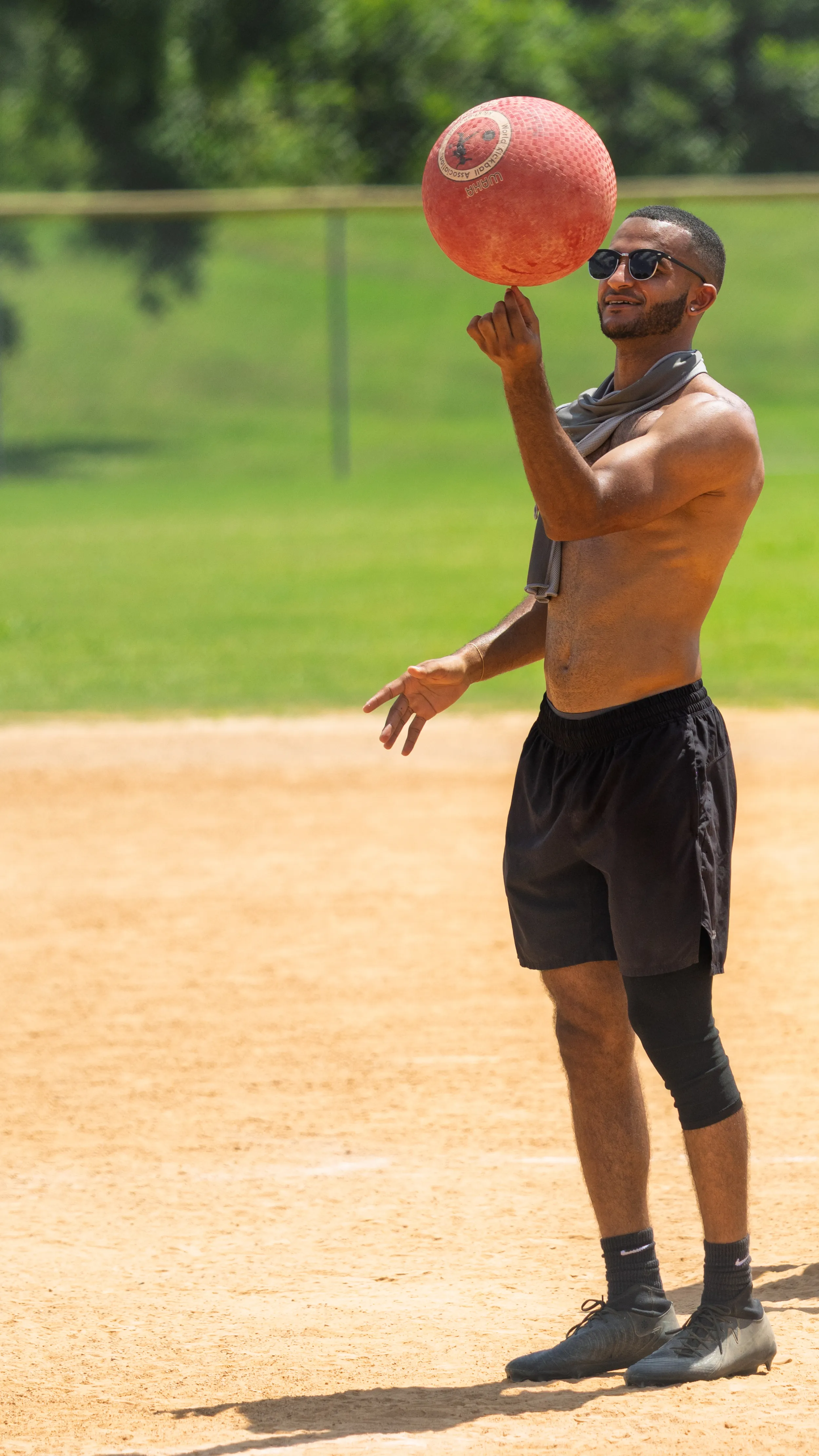 On a sunny kickball field, a player in black shorts with a knee sleeve, sunglasses, and a towel around the neck spins a red kickball on one fingertip on the dirt infield, with a fence and green outfield behind.