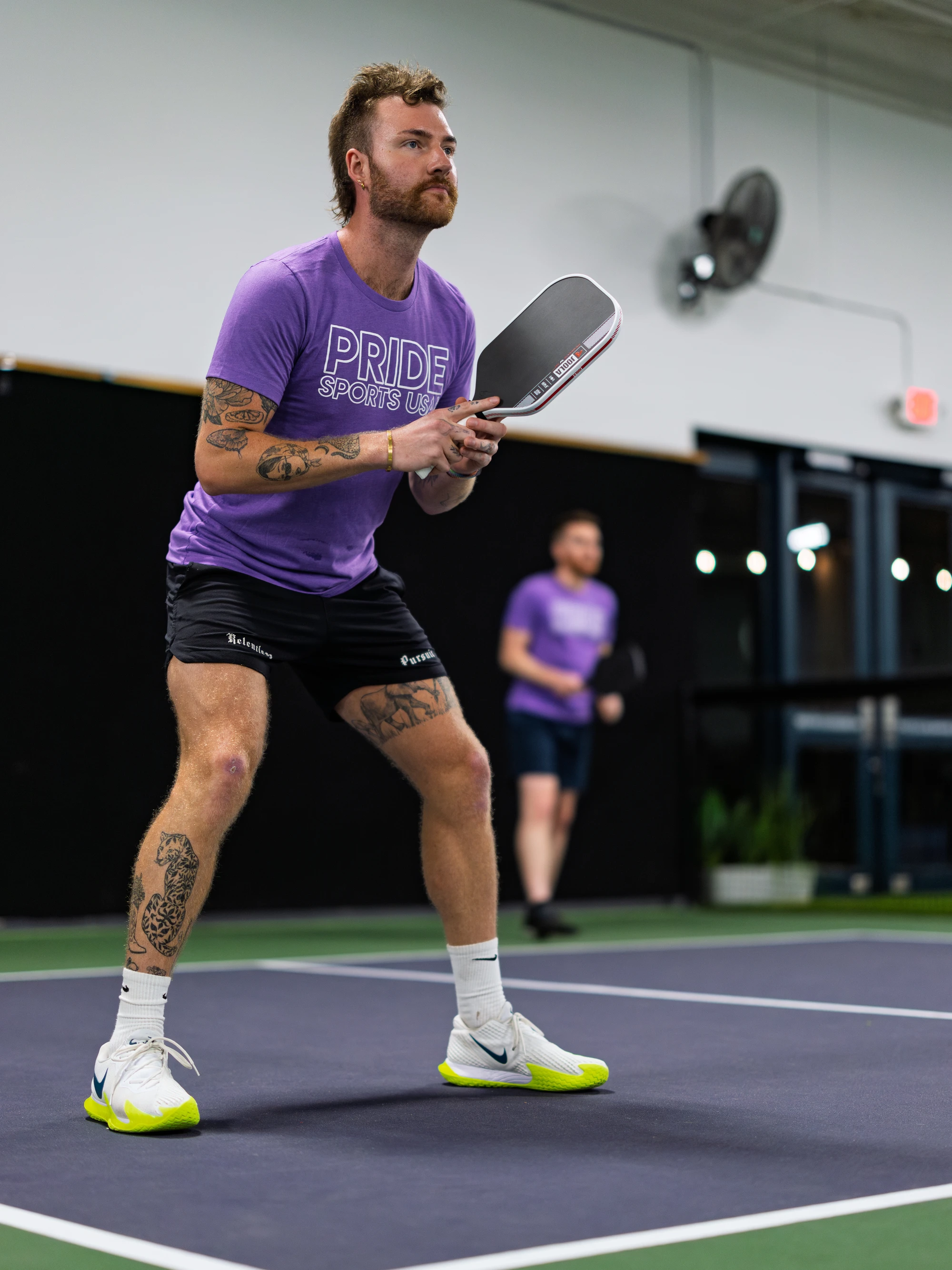 A pickleball player wearing a purple "Pride Sports USA" shirt and black shorts is positioned in an athletic stance, holding a paddle and focused on the game inside an indoor court. Another player stands in the background, also ready for action.