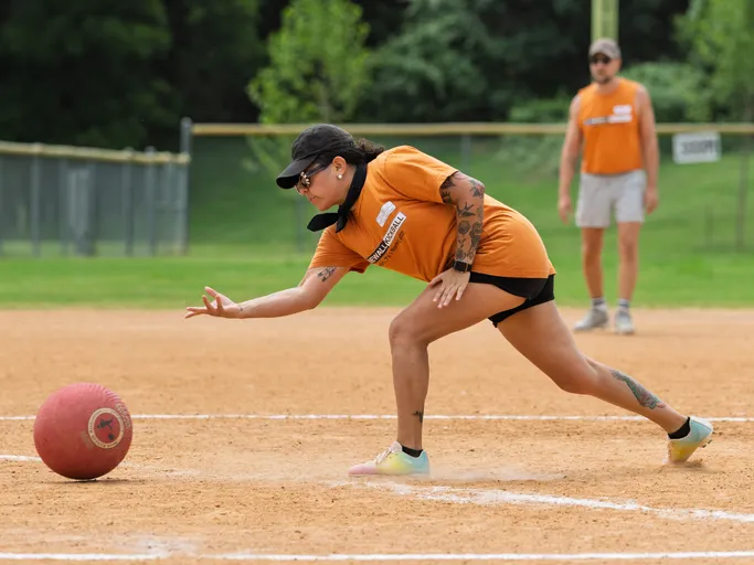 A kickball player in an orange team shirt and visor is stretching forward to pitch a red kickball on a dirt field, with another player standing in the background.
