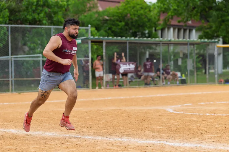A player wearing a sleeveless maroon shirt, gray athletic shorts, and red cleats is running on a kickball field with a focused expression, while teammates and spectators watch from the sideline near a chain-link fence.