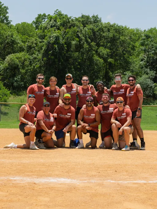 A group of kickball players pose together on a dirt field, all wearing matching red t-shirts, shorts, and various athletic shoes; some have sunglasses, hats, or headbands, with trees and green grass visible in the background.