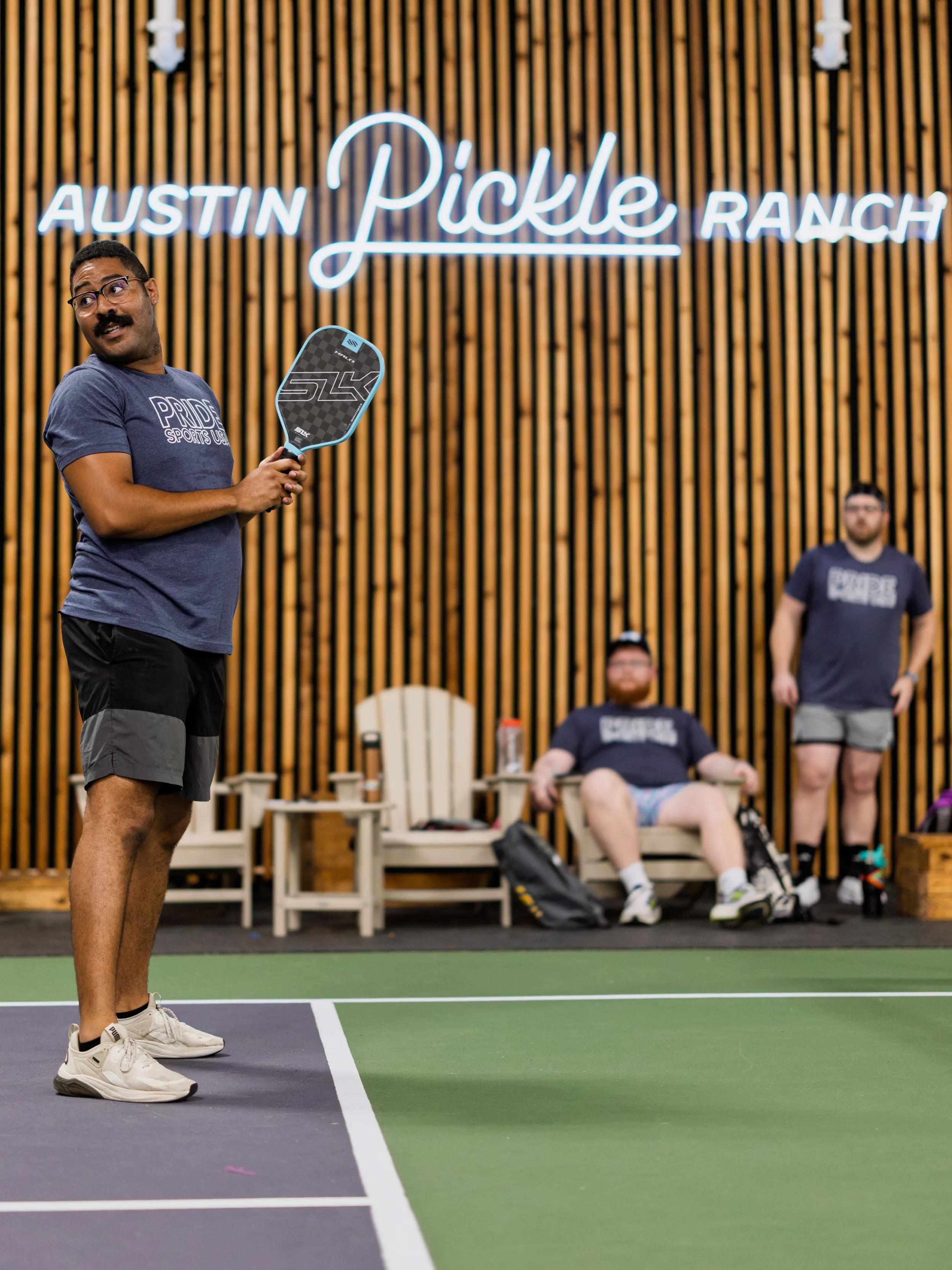 On an indoor court at Austin Pickle Ranch, a pickleball player in a navy Pride Sports League shirt, shorts, and sneakers stands at the baseline with paddle raised, looking ready to return a shot. Two players in matching shirts watch from wooden benches along the back wall beneath a neon “Austin Pickle Ranch” sign, with gear bags at their feet.