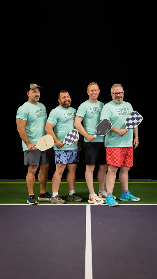 Four players stand side by side on a pickleball court, each holding a paddle and wearing matching light blue "Pride Pickleball" shirts paired with colorful shorts and athletic shoes. The group is posing together, ready for action under bright lighting, with a dark background highlighting their team spirit.