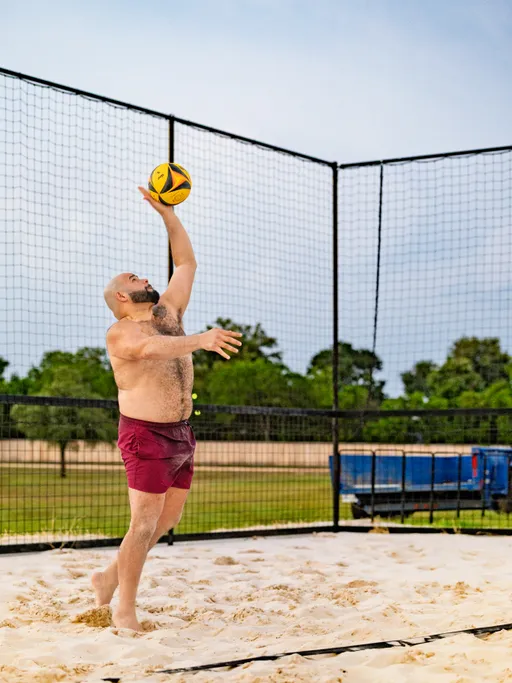 A volleyball player in maroon shorts is jumping and reaching up to hit a yellow and black volleyball on a sand court, surrounded by netting with trees and a blue trailer in the background.