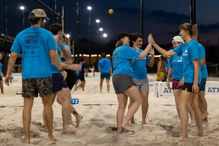 A group of sand volleyball players in matching blue league shirts huddle near the net and exchange high-fives on a lit outdoor court at dusk, wearing shorts and a mix of caps and visors, with other games and a yellow ball visible in the background.