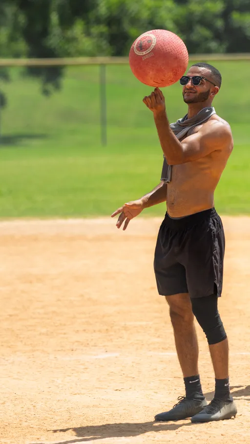 A player on a kickball field balances a red kickball on one finger while standing on the dirt, dressed in black athletic shorts, socks, sneakers, sunglasses, and a towel draped around their neck.