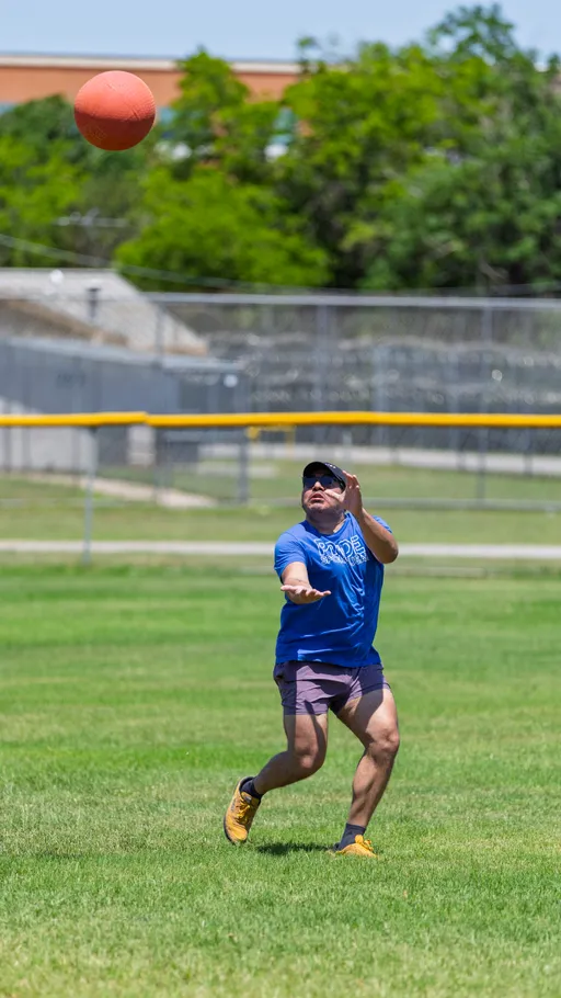 A kickball player in a blue shirt, shorts, and yellow shoes is preparing to catch a kickball in mid-air on a grassy field, with trees and a fence in the background.