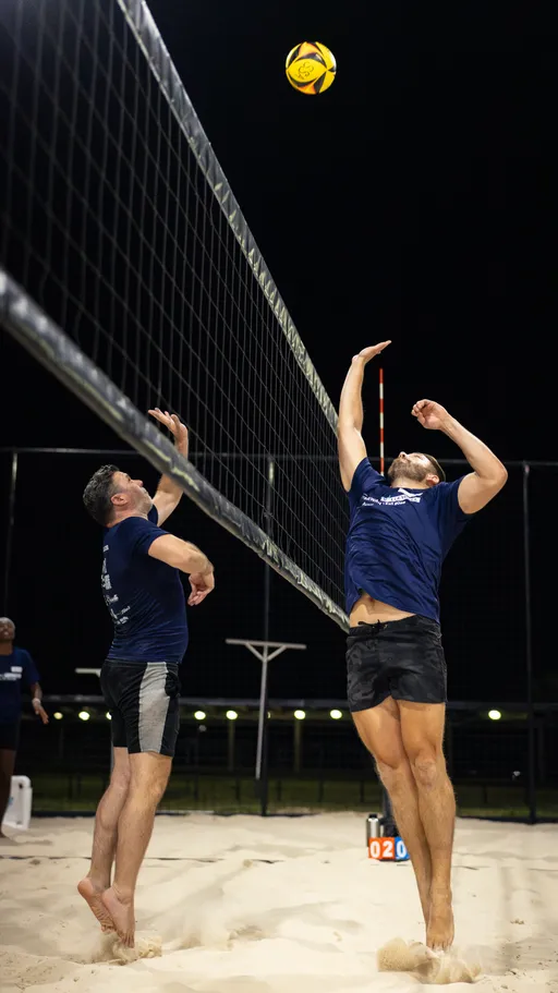 Under night lights on a sand volleyball court, two players meet at the net as a yellow-and-black ball hovers overhead; one leaps with an outstretched arm for a tip or spike while the other reaches up to block. Both wear navy shirts and dark shorts with sand kicking up beneath them, and a small scoreboard and fencing are visible in the background.