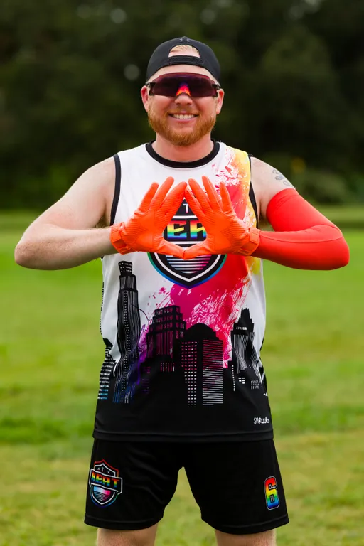 On a grassy practice field, an ATX Heat (Orange) player poses for Gay Bowl 2025 prep, wearing a black cap, mirrored sport shades, a sleeveless jersey with a colorful cityscape and team crest, black shorts with a crest and “6,” and bright orange receiver gloves and arm sleeve, holding gloved hands in a diamond shape.
