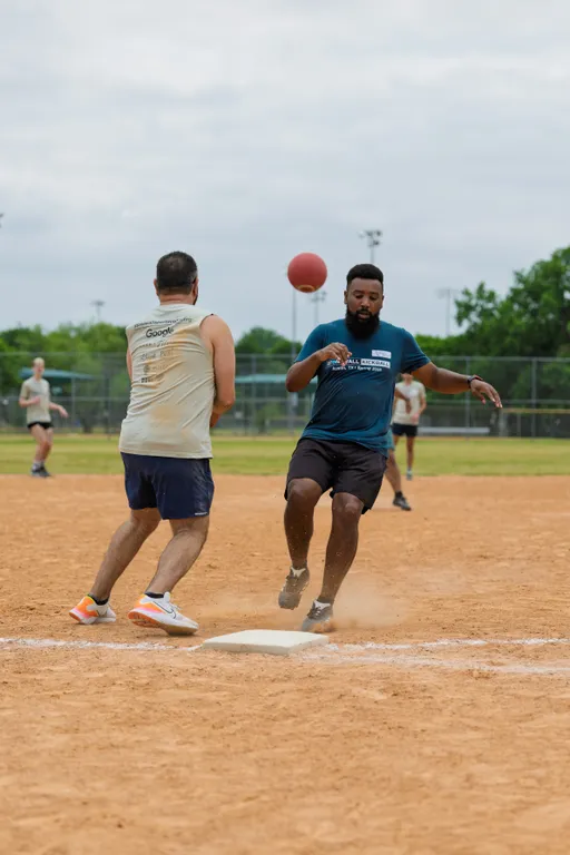 Two kickball players converge at a base, one attempting to step on the base while the other moves to intercept, as a red kickball is airborne between them on a dusty kickball field with other players visible in the background.