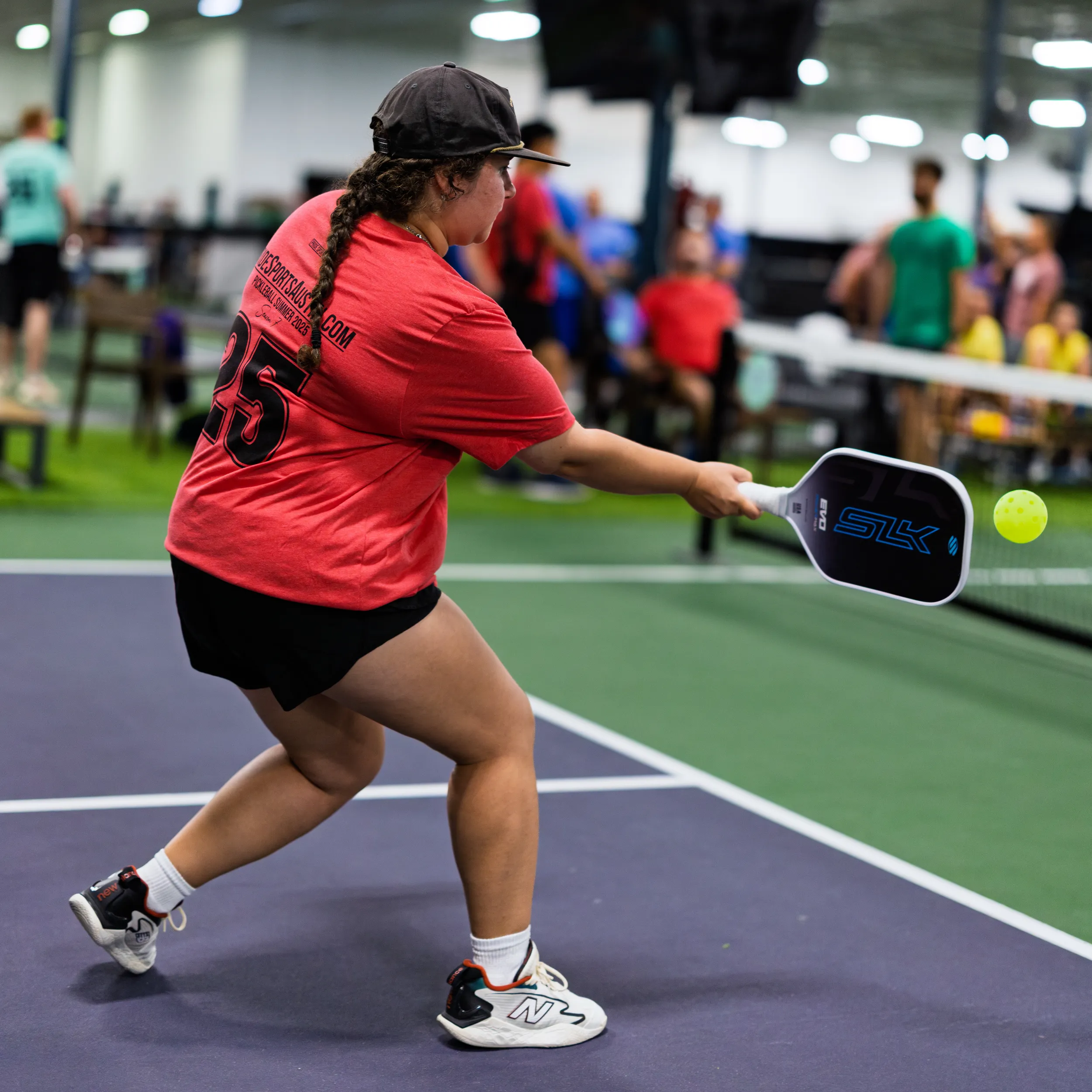 On an indoor pickleball court, a player in a red jersey with the number 25, black shorts, and a dark cap lunges to return a bright yellow pickleball with a dark paddle. Benches, other players, and spectators blur in the background, suggesting an active Pride Pickleball match in a busy multi-court facility.