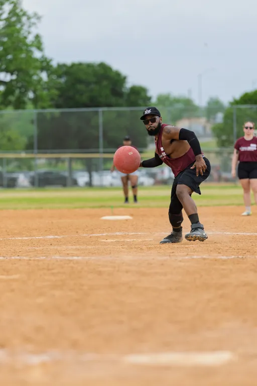 A kickball player in athletic clothing, including a tank top, shorts, cap, and arm sleeves, is mid-action on a dirt field, preparing to pitch the ball; other players and a backstop fence are visible in the background.