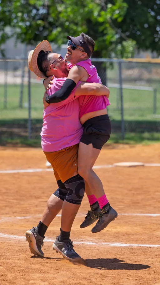 Two kickball players wearing matching pink shirts celebrate on the field, with one player lifting the other off the ground; both are wearing athletic shoes and sunglasses, and the player being lifted also has a black visor and bright pink socks. The scene takes place on a dusty outdoor kickball field with green trees and a chain-link fence in the background.