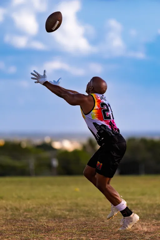 An ATX Heat (Black) player sprints forward with arms outstretched, attempting a catch on a spiraling football during Gay Bowl practice on a grassy field; wearing a rainbow-gradient sleeveless team jersey, black shorts, white receiver gloves, light cleats, and a waist belt, with blue sky and distant trees in the background.