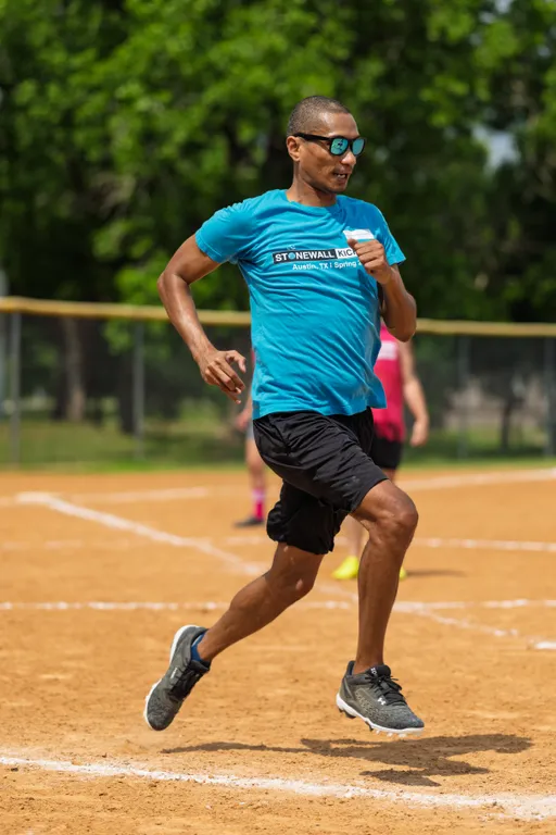 A player wearing a blue "Stonewall Kickball" shirt, black shorts, and sunglasses is running on a kickball field, with another player visible in the background.