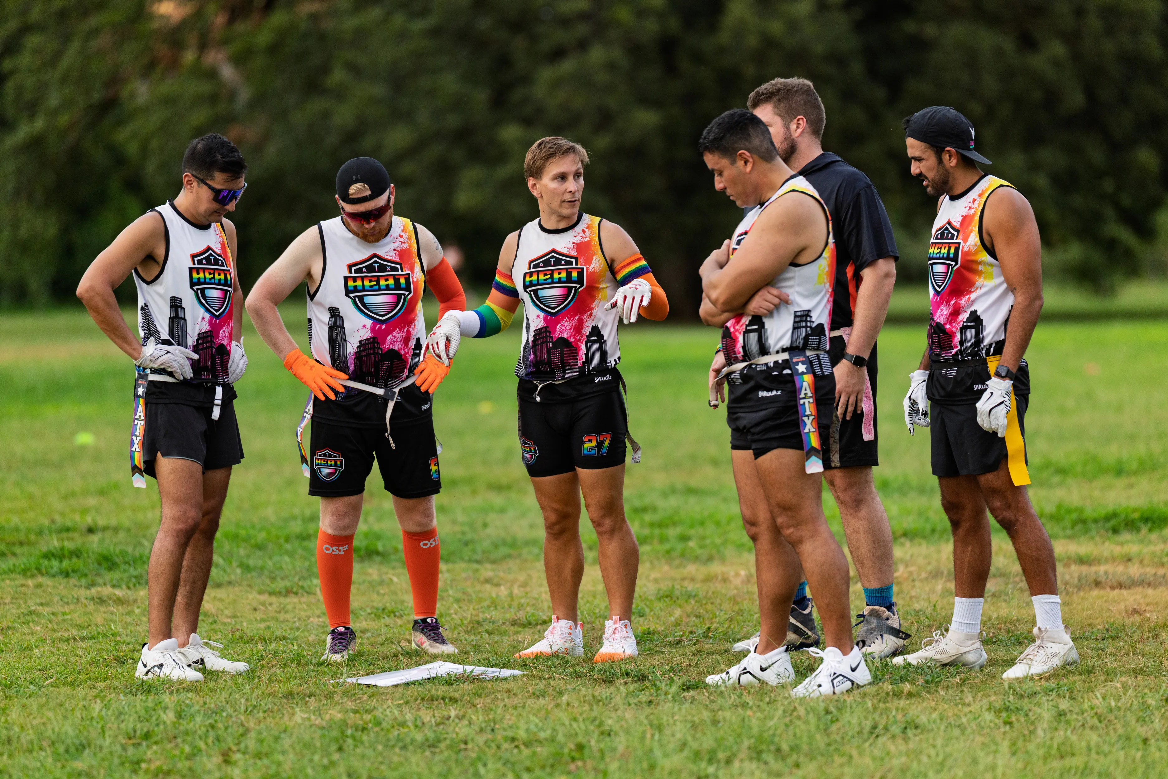 On a grassy practice field, ATX Heat (Orange) players huddle around a play sheet on the ground, discussing strategy during Gay Bowl 2025 prep. They wear matching white jerseys with the HEAT logo and orange/pink graphics, black shorts with flag belts, cleats, and assorted accessories like gloves, a cap, and an armband, with trees lining the background.