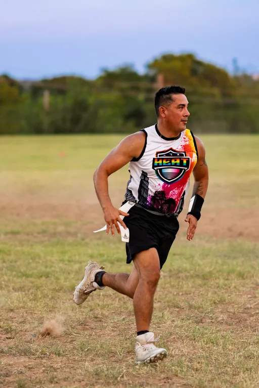 An ATX Heat (Black) flag football player sprints across a grassy practice field during Gay Bowl 2025 prep, mid-turn with eyes upfield and dust kicking up, wearing a sleeveless Heat jersey with a colorful skyline logo, black shorts with a white flag belt, cleats, and a wristband, with trees and open sky in the background.