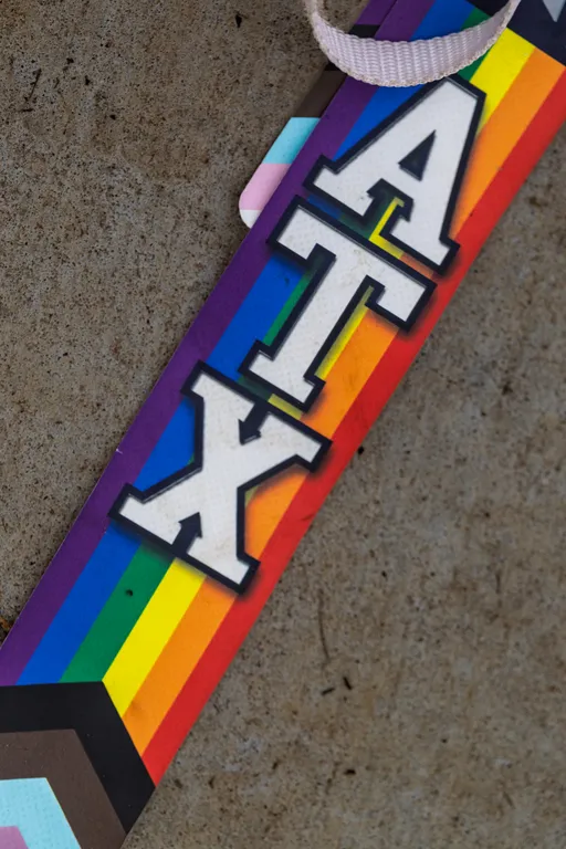 Close-up of a rainbow-striped strip on concrete with bold white letters “ATX” stacked vertically; a small white fabric loop is visible at the top, and the lower edge includes the Progress Pride chevron colors.