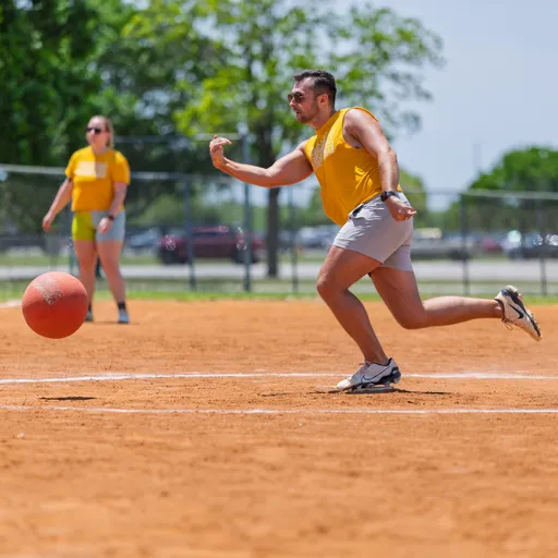 A player wearing a yellow shirt and gray shorts is in mid-action, preparing to kick a red kickball on a dirt field while another player stands in the background, also wearing a yellow shirt.