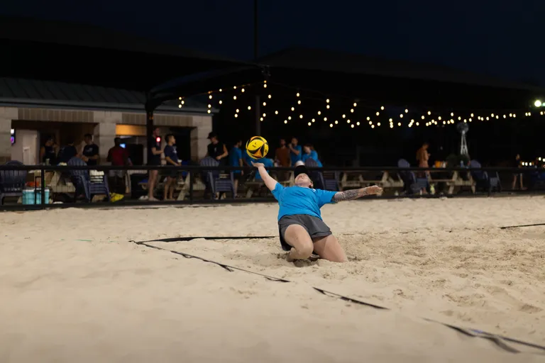 During a night match, a player in a blue shirt and gray shorts slides backward in the sand, reaching up with one hand to keep a yellow-and-black volleyball in play. String lights glow over the sand court as teammates and spectators watch from a pavilion and along the fence in the background.