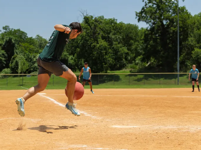A player in a dark shirt and shorts is captured mid-jump, about to kick a red kickball on a sandy field, while other players in athletic gear watch from the outfield. Trees and a fence line the background under a clear sky.