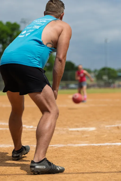 A player in a blue tank top and black shorts is ready in a crouched position on a kickball field, watching a rolling kickball being pitched by another player in the background.