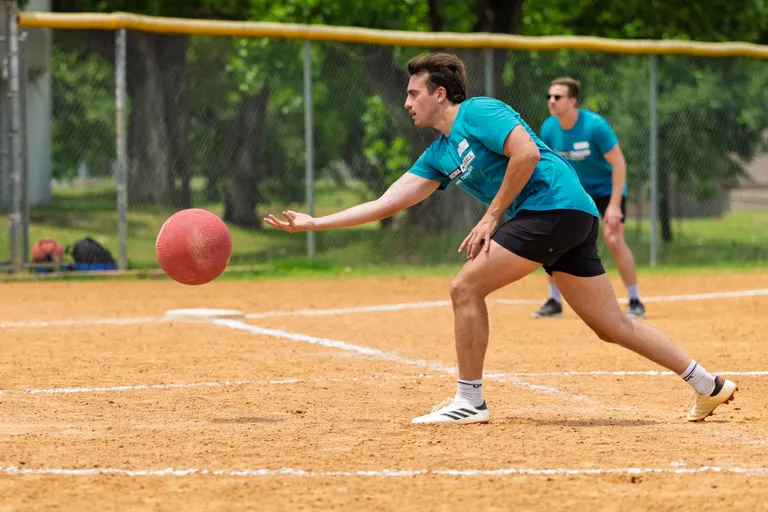 A player wearing a teal team shirt, black shorts, and athletic shoes is in the process of pitching a red kickball on a dirt field, while another player in a matching shirt and shorts stands in the outfield near a chain-link fence.
