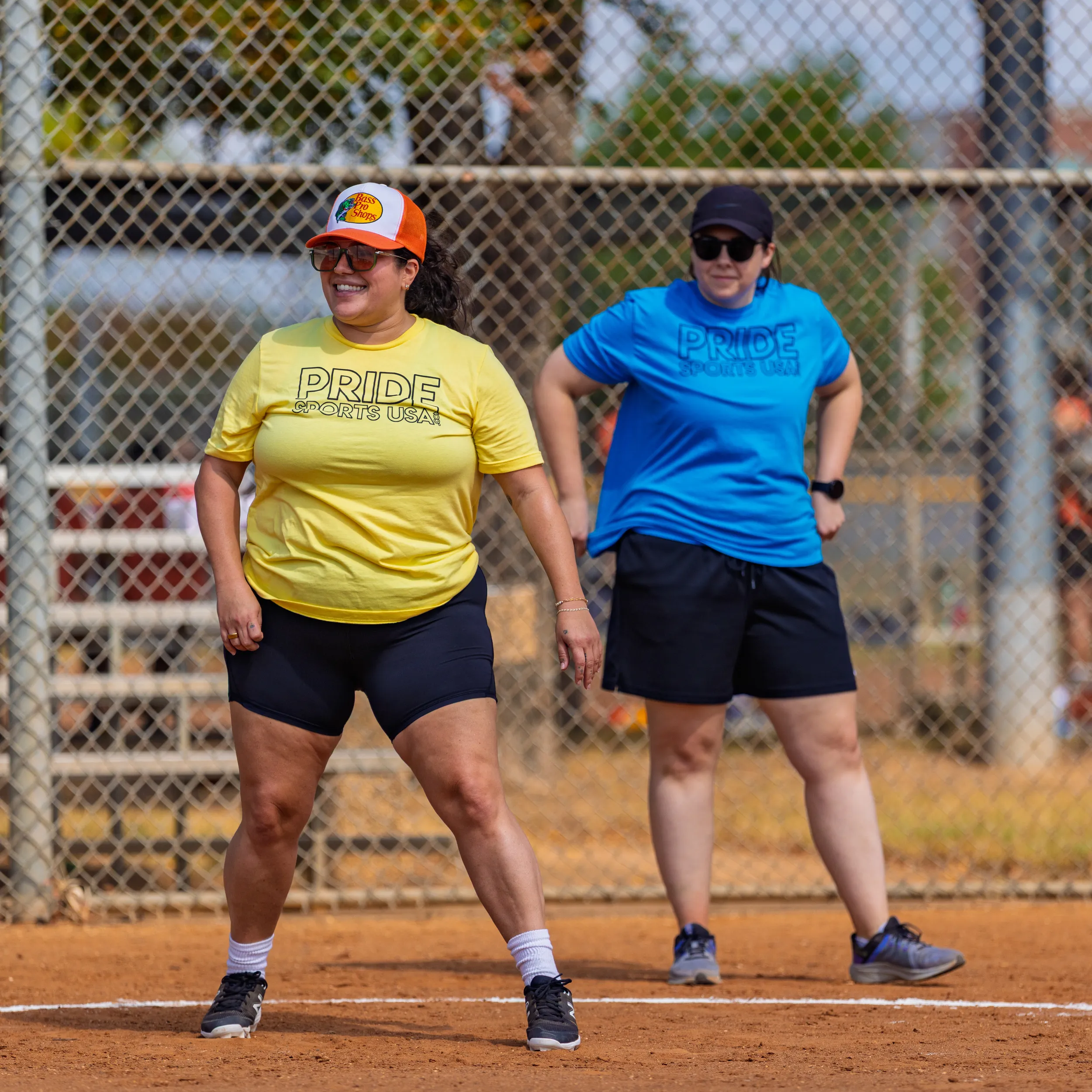 Two Pitch Please players stand on the kickball infield near a chain-link fence, pausing between plays. One wears a yellow “PRIDE” shirt, black shorts, and an orange cap; the other wears a blue “PRIDE” shirt, black shorts, and a black cap, both in athletic shoes.