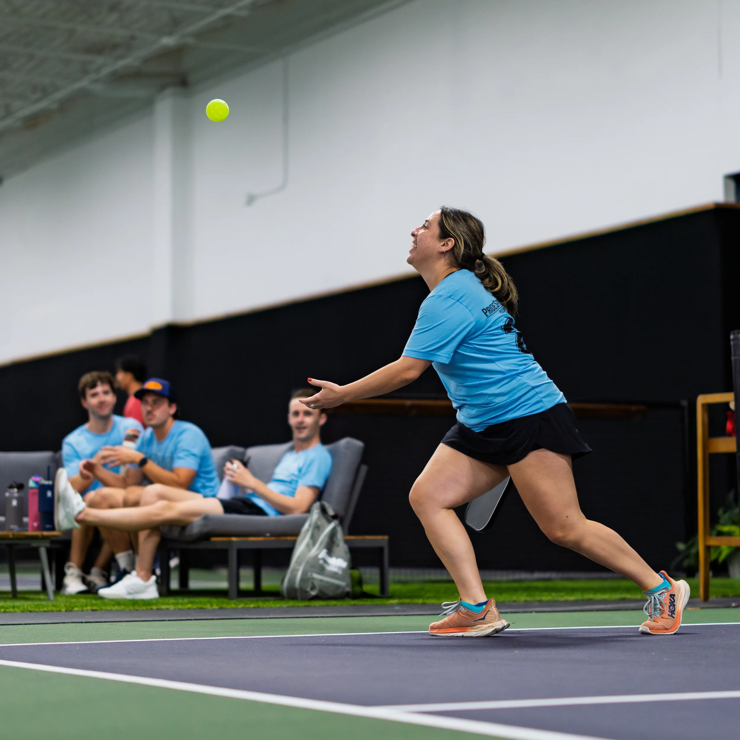 On an indoor pickleball court, a player in a light-blue team shirt, black shorts, and bright sneakers tracks a high lob and prepares to return it. Several teammates in matching shirts watch from a courtside bench with bags and water bottles beside them, set against a turf border and high ceiling.