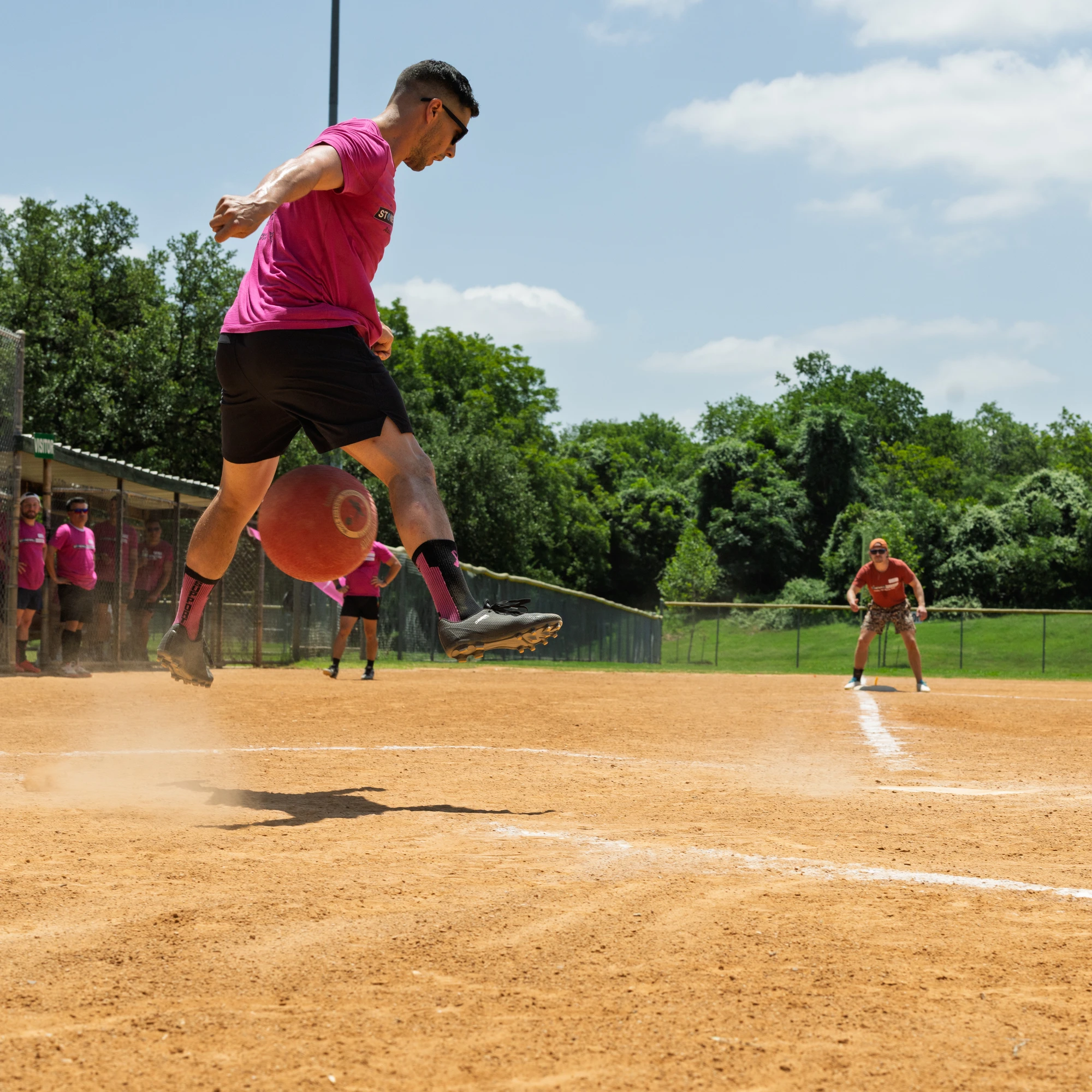 A player wearing a bright pink shirt and black shorts is mid-air after kicking a red kickball on a dusty field, while other athletes stand ready in the background near the outfield and dugout.
