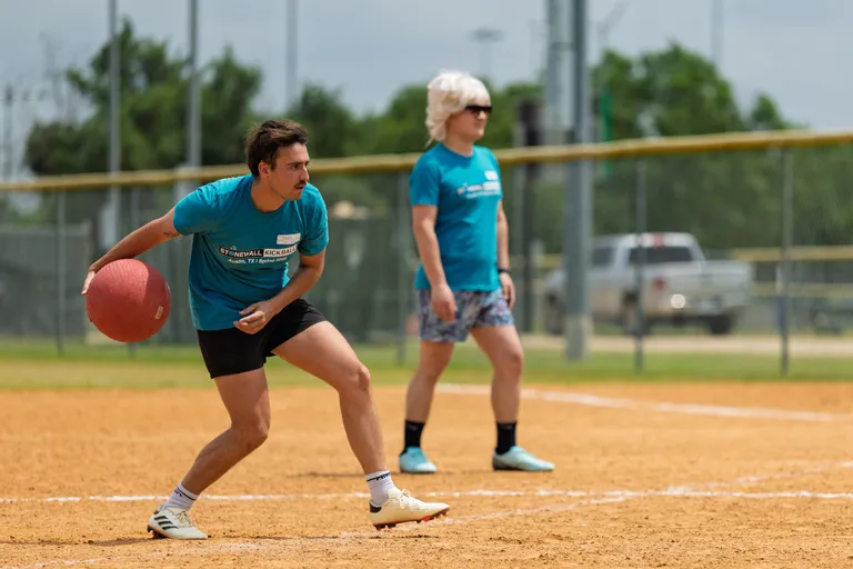 Two players on a kickball field are wearing turquoise team shirts; one is in a ready position holding a red kickball, preparing to throw or pitch, while the other stands nearby watching the play.