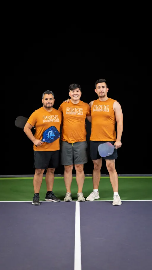 Three pickleball players stand together on an indoor court, each wearing matching orange "Pride Sports USA" shirts and holding paddles, posing in a celebratory team photo.