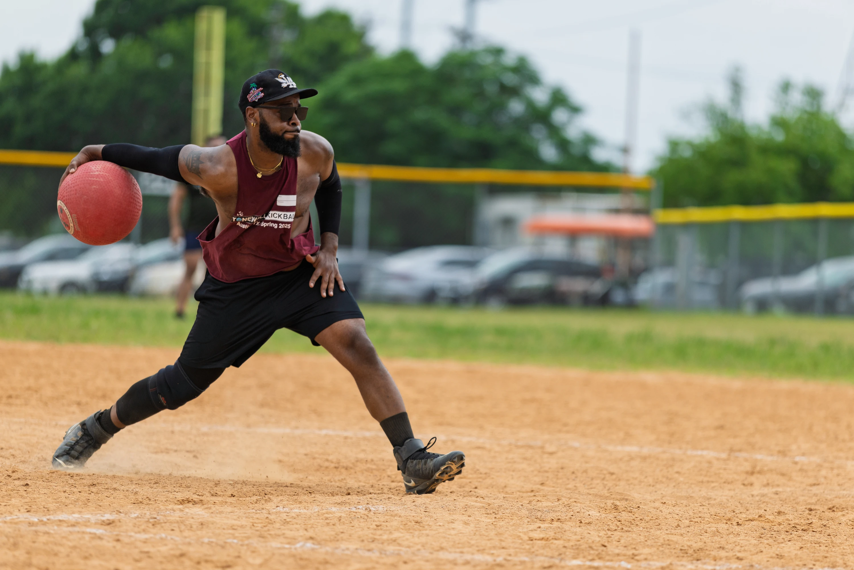 A kickball player wearing a maroon jersey, black athletic gear, and a cap is in mid-action, preparing to throw a red kickball on a sandy field with a fence and parked cars in the background.