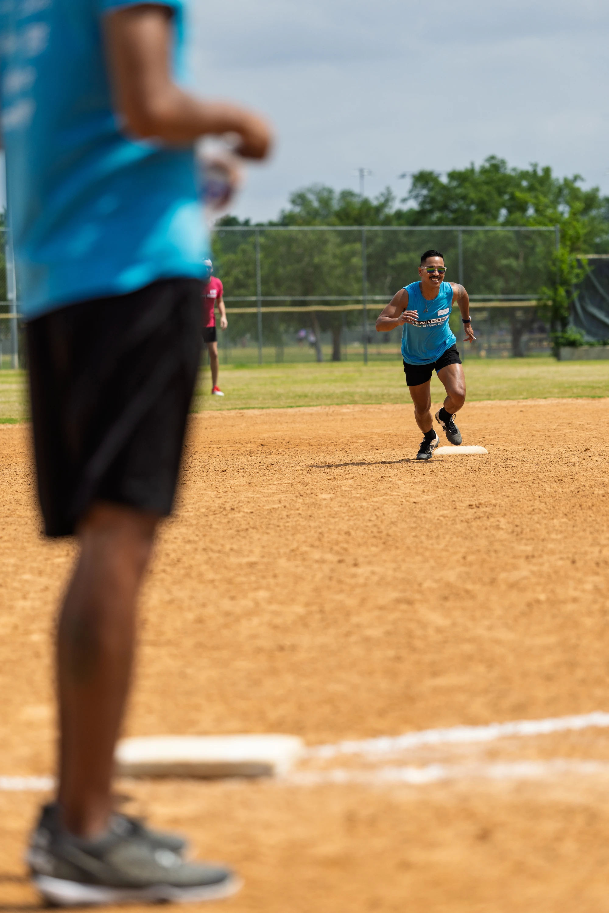 A kickball player in a blue tank top and black shorts is running from a base on a dirt kickball field, while another player stands in the foreground wearing similar athletic clothing and sneakers. A chain-link fence and grassy area are visible in the background.