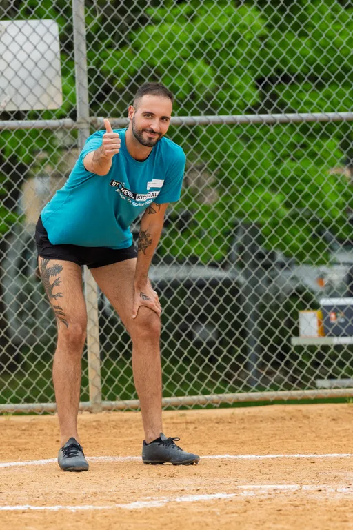 A player stands on a kickball field near home plate, wearing a teal Stonewall Kickball shirt, black shorts, and athletic shoes, smiling and giving a thumbs-up gesture while leaning forward with one hand on their knee. The background features a chain-link fence and green foliage.
