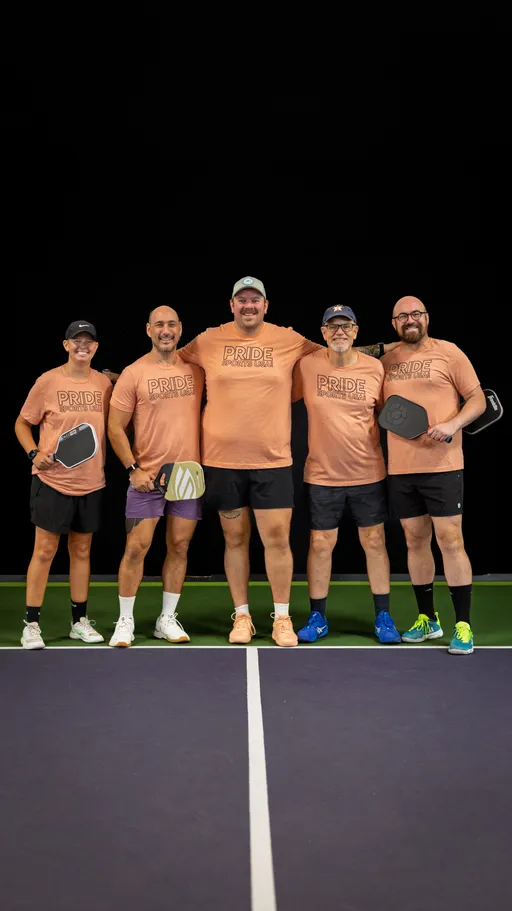 Five pickleball players stand arm in arm on an indoor court, each wearing matching "Pride Pickleball" t-shirts, shorts, and athletic shoes, while holding their pickleball paddles and smiling towards the camera.