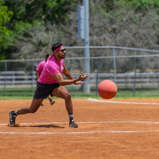 A player wearing a pink shirt, black shorts, and a pink headband is reaching out with both hands to catch a red kickball on a dirt kickball field, with a fence and trees visible in the background.