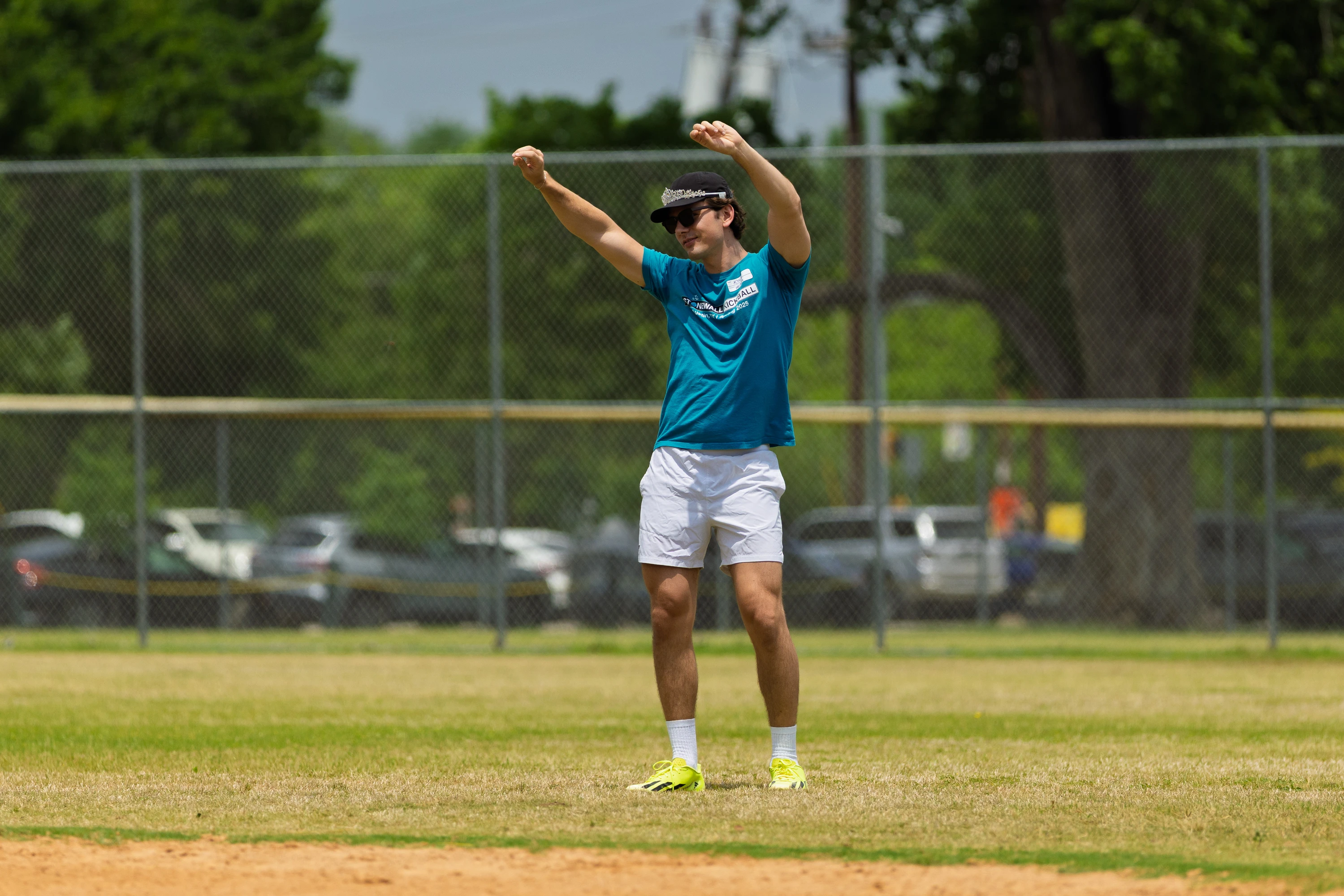 A player in a teal shirt, white shorts, bright shoes, and a visor is standing on the kickball field with both arms raised, possibly celebrating or signaling during a game. The scene takes place outdoors with a chain-link fence and trees in the background.