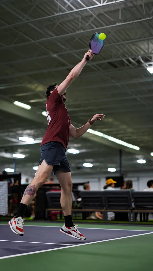 A pickleball player in athletic clothing jumps to hit a ball with a paddle on an indoor court, with spectators seated in the background.